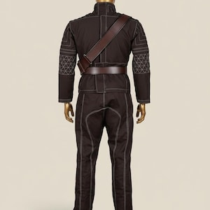 Puede incluir: Mono marr&oacute;n con costuras blancas, bandolera y cintur&oacute;n de cuero marr&oacute;n, expuesto en un maniqu&iacute;. El conjunto presenta un cuello alto y patrones geom&eacute;tricos en las mangas. El maniqu&iacute; tiene cabeza y manos doradas.