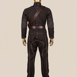 Puede incluir: Mono marr&oacute;n con costuras blancas y patrones geom&eacute;tricos en los hombros y las mangas. El atuendo incluye un cintur&oacute;n marr&oacute;n y una correa para el hombro. El maniqu&iacute; tiene cabeza y manos doradas.