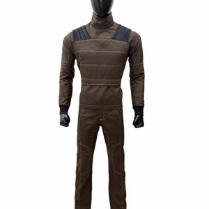 May include: A brown jumpsuit displayed on a mannequin. The outfit includes a long-sleeved top with a high collar and horizontal stitching, and matching pants. The shoulders have dark gray accents, and the cuffs are black.