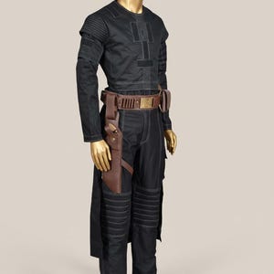 May include: Black costume with white stitching, displayed on a mannequin. The outfit includes a long-sleeved top, pants, and a brown leather belt with a holster. The pants have padded knee details. The mannequin has a gold finish.
