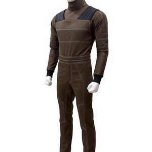 May include: Brown jumpsuit with white stitching details, displayed on a mannequin. The jumpsuit features a high collar, long sleeves with black cuffs, and a tailored fit. The design includes shoulder straps and horizontal stripes across the chest.