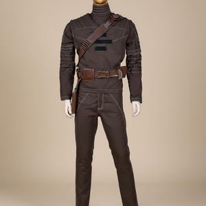 Puede incluir: Un disfraz marr&oacute;n exhibido en un maniqu&iacute;. El atuendo incluye un top de cuello alto, pantalones y un cintur&oacute;n con una funda. Las costuras blancas acent&uacute;an las costuras. Una correa de hombro marr&oacute;n y un cintur&oacute;n con una bolsa completan el look. El maniqu&iacute; tiene una cabeza dorada.