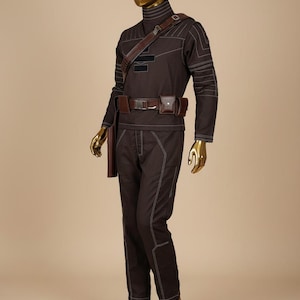 Puede incluir: Un disfraz marr&oacute;n con costuras blancas, exhibido en un maniqu&iacute; con cabeza y manos doradas. El atuendo incluye una parte superior de manga larga, pantalones, un cintur&oacute;n con bolsillos y una correa para el hombro. El disfraz est&aacute; dise&ntilde;ado con un cuello alto.