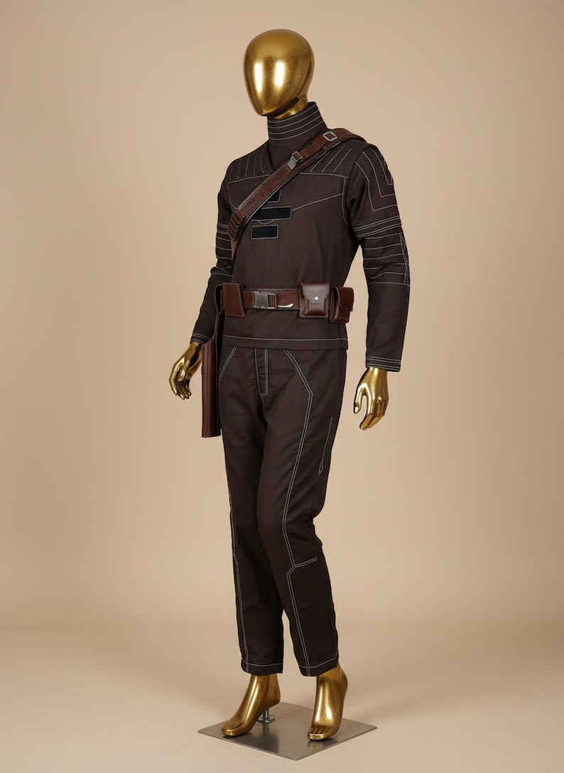 Puede incluir: Un disfraz marr&oacute;n con costuras blancas, exhibido en un maniqu&iacute; con cabeza y manos doradas. El atuendo incluye una parte superior de manga larga, pantalones, un cintur&oacute;n con bolsillos y una correa para el hombro. El disfraz est&aacute; dise&ntilde;ado con un cuello alto.