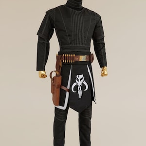 May include: A black costume with white stitching, featuring a high collar and long sleeves. The outfit includes a brown belt with ammunition, a holster, and a black apron with a white emblem. The mannequin has a gold head and feet.
