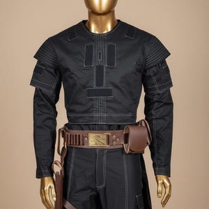 May include: Black costume with white stitching, featuring a long-sleeved top with shoulder accents and a matching pair of pants. A brown leather belt with a rectangular buckle and a pouch is worn around the waist. A holster is attached to the belt.