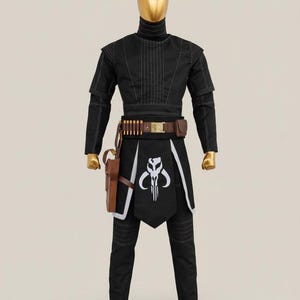 May include: Black costume with white stitching, featuring a high collar and a distinctive emblem on the front. The outfit includes a brown belt with ammunition, a holster, and matching trousers. The mannequin has a gold head and feet.