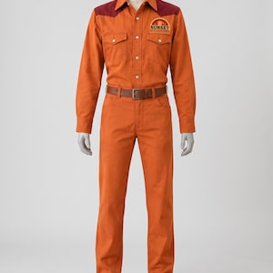 Op de afbeelding: Een oranje overhemd met lange mouwen, knoopsluiting, een donkerrood schouderstuk en het Sunset Sarsaparilla-logo op de borst, gecombineerd met een bijpassende oranje broek, een bruine leren riem en bruine leren laarzen op een etalagepop.