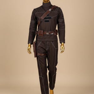 Puede incluir: Un disfraz marr&oacute;n con costuras blancas, que incluye una parte superior de manga larga y pantalones. El atuendo incluye un cintur&oacute;n marr&oacute;n, una correa para el hombro y una funda. El maniqu&iacute; tiene cabeza y manos doradas.