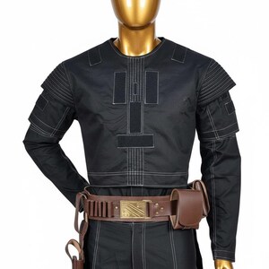 May include: Black costume with white stitching details, displayed on a mannequin. The outfit includes a long-sleeved top, pants, and a brown leather belt with a rectangular buckle. A holster and pouch are also included.