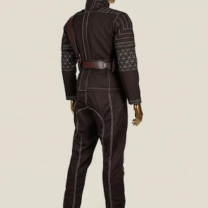 Puede incluir: Mono marr&oacute;n oscuro con costuras blancas y patrones geom&eacute;tricos en las mangas. El atuendo incluye un cintur&oacute;n marr&oacute;n y una correa para el hombro. El maniqu&iacute; tiene cabeza y manos doradas.