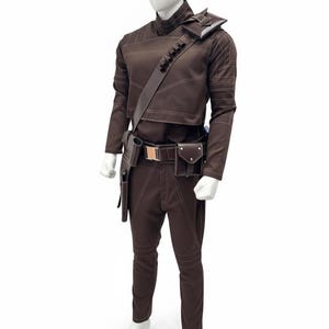 May include: A brown costume displayed on a mannequin. The outfit includes a long-sleeved top, pants, and a belt with pouches. White stitching accents the seams. A shoulder strap and holster are also included. The mannequin is on a white background.