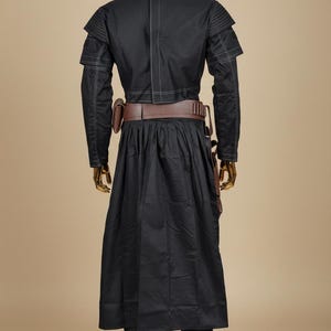 May include: Black costume with a long coat, pants, and a brown belt with pouches. The costume has white stitching details and is displayed on a gold mannequin. The outfit is reminiscent of a sci-fi or fantasy character.