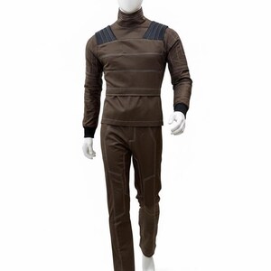 May include: A brown jumpsuit displayed on a mannequin. The outfit features a high collar, long sleeves, and pants. White stitching outlines the seams and panels. Black accents are on the shoulders and wrists. The mannequin is white, and the background is plain.
