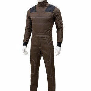 May include: Brown jumpsuit with white stitching details, displayed on a mannequin. The jumpsuit features a high collar, black shoulder accents, and black cuffs. The garment has a utilitarian design, with visible seams and reinforced panels.