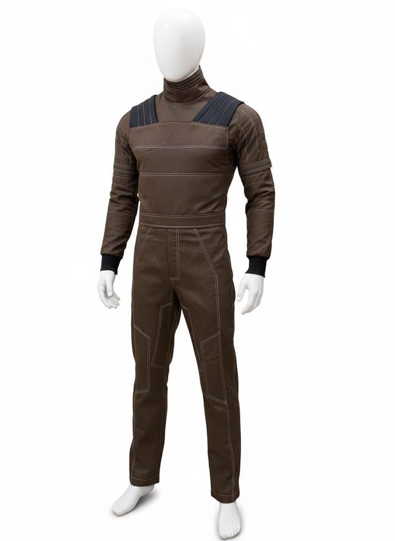 May include: Brown jumpsuit with white stitching details, displayed on a mannequin. The jumpsuit features a high collar, black shoulder accents, and black cuffs. The garment has a utilitarian design, with visible seams and reinforced panels.
