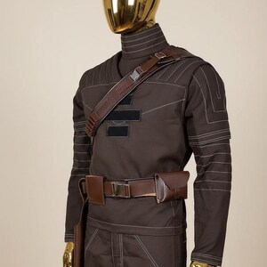 Puede incluir: Un disfraz marr&oacute;n con detalles de costura blancos, con una parte superior de cuello alto, pantalones y un cintur&oacute;n de cuero marr&oacute;n con bolsas. El disfraz incluye una correa para el hombro y se muestra en un maniqu&iacute; con una cabeza dorada.