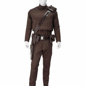 May include: Brown costume with a high collar, long sleeves, and visible stitching. The outfit includes matching pants, a belt with pouches, and a shoulder strap with a rectangular accent. The mannequin is on a silver base.