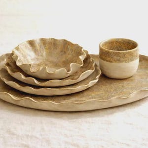 Pode incluir: Um conjunto de louça de cerâmica composto por uma travessa oval grande, dois pratos empilhados, uma tigela pequena e uma chávena. Cada peça tem uma borda ondulada e festonada com um acabamento texturizado em tons de bege e castanho.