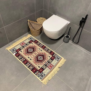 May include: A rectangular bathroom rug with a cream border and fringe. The rug has a geometric pattern in red, black, and beige. A white toilet, a toilet brush, and a woven basket are in the background.