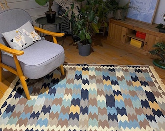 Op de afbeelding: Een rechthoekig vloerkleed met een zigzagpatroon in blauw, bruin en beige tinten. Het kleed heeft een crèmekleurige franje. Het kleed ligt op een houten vloer. Afmetingen: ongeveer 200 cm x 140 cm.