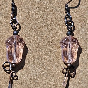 以下が含まれることがあります： 淡いピンク色のチューリップ型のガラスビーズが特徴的な、手作りのイヤリングのペア。イヤリングは、装飾的なループと涙型のドロップが付いた、暗いアンティーク調の金属デザインです。イヤーフックはシンプルなフックタイプです。
