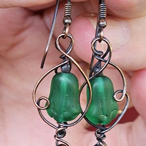以下が含まれることがあります： 緑色のベル型のガラスビーズを使用した手作りのイヤリング。イヤリングは、渦巻きやループを含む複雑な銅線細工が施されています。 スタイルは繊細で芸術的で、ヴィンテージ感があります。