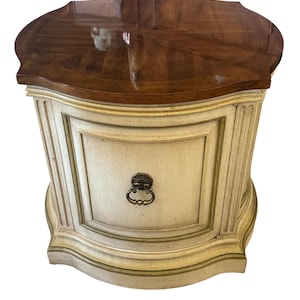 May include: A small, antique-style nightstand with a curved, dark brown wooden top. The base is off-white with gold trim and a decorative drawer pull. The nightstand has a classic, elegant design, suitable for a bedroom.