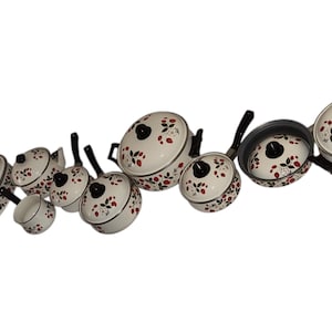 May include: A collection of white enamel cookware with black handles and lids. The cookware is decorated with a red and black floral pattern. The set includes various pots, pans, and lids, suitable for cooking a variety of dishes.