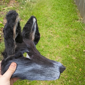 以下が含まれることがあります： 黒とグレーのフェイクファーで作られた動物のマスク。高い尖った耳にはライトグレーのアクセントがあり、長い鼻先と暗い三角形の鼻が付いています。マスクは質感のある毛むくじゃらの外観で、黒い毛からグレーの毛へとグラデーションになっています。