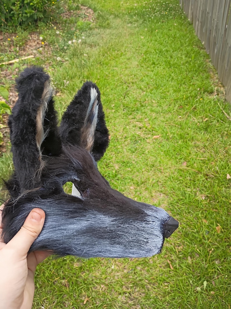 以下が含まれることがあります： 黒とグレーのフェイクファーで作られた動物のマスク。高い尖った耳にはライトグレーのアクセントがあり、長い鼻先と暗い三角形の鼻が付いています。マスクは質感のある毛むくじゃらの外観で、黒い毛からグレーの毛へとグラデーションになっています。