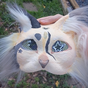 以下が含まれることがあります： クリーム色のベースに黒と灰色のペイントが施された手作りの猫のマスク。耳と側面には白と灰色の長いふわふわしたフェイクファーの房があり、額には小さなハートの模様、目にはメッシュのカバーが付いています。