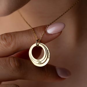 May include: A gold-colored pendant necklace featuring three interlocking circles. Each circle has a name engraved on it. The necklace is suspended from a delicate chain. The background is a blurred view of a person's skin.