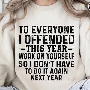 May include: Cream-colored sweatshirt with the text "TO EVERYONE I OFFENDED THIS YEAR WORK ON YOURSELF SO I DON'T HAVE TO DO IT AGAIN NEXT YEAR" in bold black letters. The sweatshirt has a crew neck and long sleeves.
