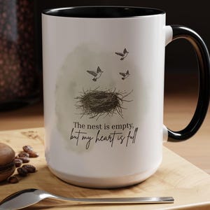 Puede incluir: Taza de cerámica blanca con interior y asa negros. La taza presenta una ilustración de nido y pájaros con el texto "The nest is empty, but my heart is full."