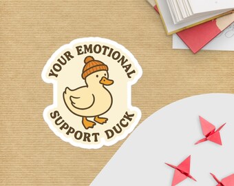 May include: A sticker featuring a cartoon duck wearing a knit cap. The text on the sticker reads "Your Emotional Support Duck". The duck is light yellow with an orange beak and feet. The sticker is on a tan surface.