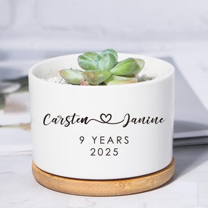 Puede incluir: Una maceta de cerámica blanca con una planta suculenta, sobre una base de madera. La maceta está inscrita con los nombres "Carsten & Janine", "9 YEARS" y "2025". La suculenta tiene hojas verdes.