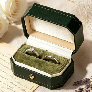 May include: An open, dark green, octagonal ring box with a gold clasp, holding two rings on a green velvet interior. One ring has a diamond center stone and smaller stones around it. The other ring is a simple band.