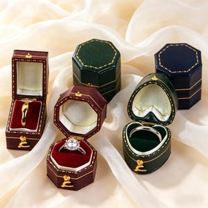 May include: An assortment of open and closed jewelry boxes in various colors, including burgundy, green, and navy blue. The boxes are octagonal and heart-shaped, with gold accents and red or green velvet interiors. Some boxes contain rings.