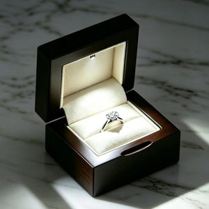 May include: A silver ring with a central cluster of small diamonds is displayed inside a dark brown wooden box. The interior of the box is lined with cream-colored fabric and features a small integrated LED light that illuminates the ring.