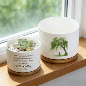 Puede incluir: Dos macetas de cerámica blanca con bases de madera. Una contiene una suculenta y pequeñas piedras blancas, con el texto "Here's to a love that bends gracefully...Happy Anniversary!". La otra presenta una ilustración de sauce llorón verde.