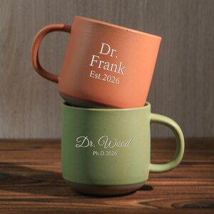 Könnte beinhalten: Zwei Keramikbecher, die auf einer Holzoberfläche gestapelt sind. Der obere Becher ist orange mit dem weißen Aufdruck "Dr. Frank Est.2026". Der untere Becher ist grün mit dem weißen Aufdruck "Dr. Wood Ph.D.2026". Beide Becher haben einen dunkelbraunen Boden und passende Henkel.