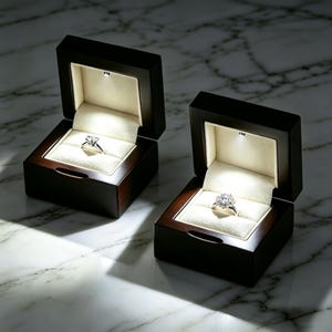 May include: Two dark brown wooden ring boxes with cream-colored velvet interiors, each containing a silver-toned ring with a clear gemstone. Each box features a small integrated LED light at the top, illuminating the rings from above.