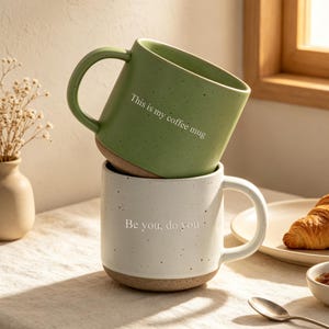 May include: A sage green speckled ceramic mug with the text This is my coffee mug stacked on top of a white speckled ceramic mug with the text Be you, do you. Both mugs feature a natural clay base and a curved handle for a simple, functional design.