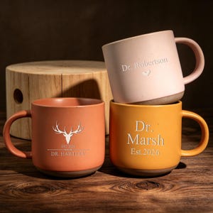 May include: Three ceramic mugs with speckled finishes in light pink, orange, and yellow. The pink mug features the text Dr. Robertson and a heart. The orange mug displays a deer antler graphic and Dr. Hartley Est. 2026. The yellow mug says Dr. Marsh Est. 2026.