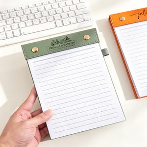 Può includere: Un blocco note verde salvia con linee orizzontali e una striscia di rilegatura superiore con due rivetti metallici dorati e il testo stampato PRISCILLA BARLOW RECORD con un motivo floreale. Un secondo blocco note arancione è visibile a destra.
