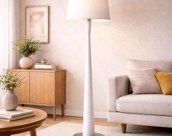 May include: A white floor lamp with a conical base and a cylindrical lampshade. The lamp has a silver-colored base and is positioned next to a beige sofa. The room also features a wooden cabinet and a round coffee table.