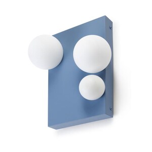 Pode incluir: Arandela de parede moderna com uma base retangular azul pó e três luminárias esféricas brancas. As luminárias são de tamanhos variados, criando um design único. A arandela é montada em uma parede branca.