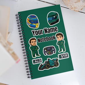 Puede incluir: Cuaderno de espiral verde con el texto "Your Name Notebook". La portada presenta pegatinas con temática de carreras, incluyendo cascos, un volante, un coche de carreras y figuras de dibujos animados con trajes de carreras verdes.