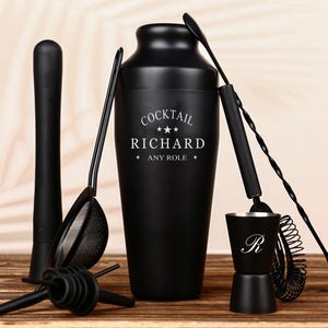 May include: A black cocktail shaker set with a variety of bar tools. The shaker is engraved with "COCKTAIL RICHARD ANY ROLE". The set includes a muddler, strainer, jigger, and bar spoon. The tools are arranged on a wooden surface.